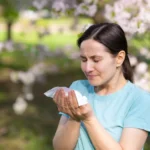 Mujer con alergia primaveral estornudando bajo árboles en flor